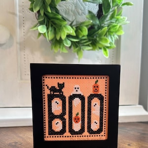 May include: A framed cross-stitch design with a peach background featuring the word "BOO" in black with white ghosts and pumpkins. A black cat sits on top of the "B".