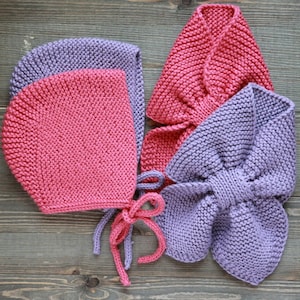 May include: Two knitted hats, one pink and one purple, with matching scarves. The hats have ties and the scarves are folded into a bow shape.