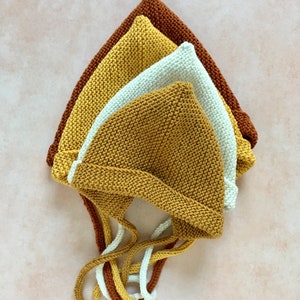 May include: Three knitted baby hats in different colors: mustard yellow, cream, and brown. The hats have long ties that can be tied under the chin.
