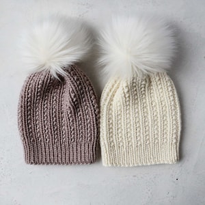 May include: Two knitted beanies with faux fur pom-poms. One beanie is a muted mauve color, and the other is off-white. The beanies have a ribbed knit pattern and a large, fluffy white pom-pom on top. The background is a light gray.