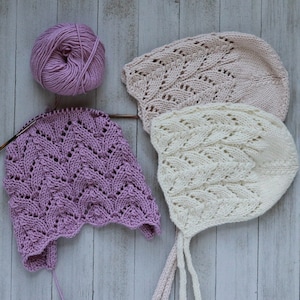 May include: Three knitted baby bonnets in pink, white, and purple with a delicate lace pattern. The bonnets are laid out on a white wooden surface.
