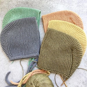 May include: Five knitted baby hats in different colors, including gray, green, yellow, and orange, with a ball of yarn and knitting needles.
