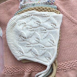 Op de afbeelding: Een witte gebreide baby muts met een delicaat bladmotief. De muts is gevouwen en gelegd op een roze gebreide trui.