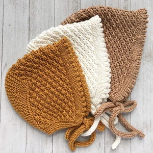 May include: Three knitted baby bonnets in different colors: mustard yellow, cream, and brown. Each bonnet has a gathered top and ties at the back.