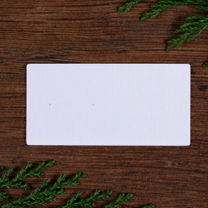 May include: A white rectangular card with two small holes, set against a dark brown wooden background. Green sprigs are placed in the corners. The card has a textured surface, possibly for displaying earrings or small jewellery.