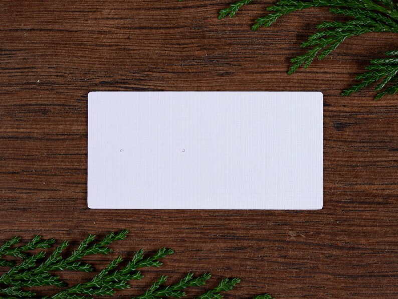 May include: A white rectangular card with two small holes, set against a dark brown wooden background. Green sprigs are placed in the corners. The card has a textured surface, possibly for displaying earrings or small jewellery.