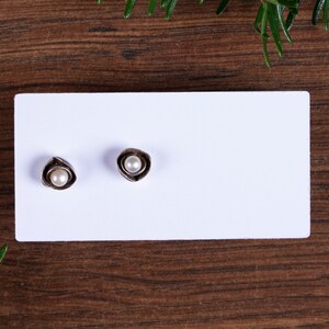 May include: A pair of silver-coloured stud earrings, each with a small, round, white pearl set within a stylized, petal-like design. The earrings are displayed on a white card against a dark wood grain background.