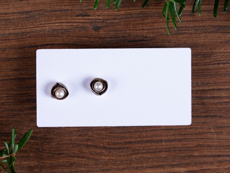 May include: A pair of silver-coloured stud earrings, each with a small, round, white pearl set within a stylized, petal-like design. The earrings are displayed on a white card against a dark wood grain background.