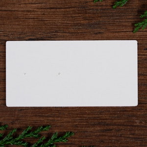 May include: A white rectangular earring display card with two small holes. The card is set against a dark wood grain background, with green sprigs in the corners.