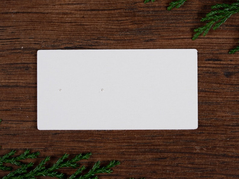 May include: A white rectangular earring display card with two small holes. The card is set against a dark wood grain background, with green sprigs in the corners.