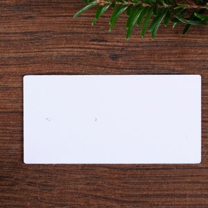 May include: A white rectangular earring display card with two small holes, set against a dark brown wooden surface. Green pine needles are in the top right and bottom left corners, adding a natural touch to the presentation.