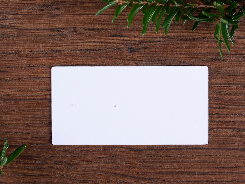 May include: A white rectangular earring display card with two small holes, set against a dark brown wooden surface. Green pine needles are in the top right and bottom left corners, adding a natural touch to the presentation.