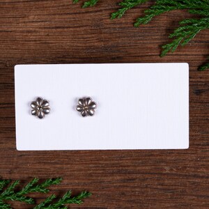 May include: A pair of flower-shaped stud earrings. Each earring has a silver-coloured, six-petal design with a gold-coloured centre. The earrings are displayed on a white card against a dark wood background.