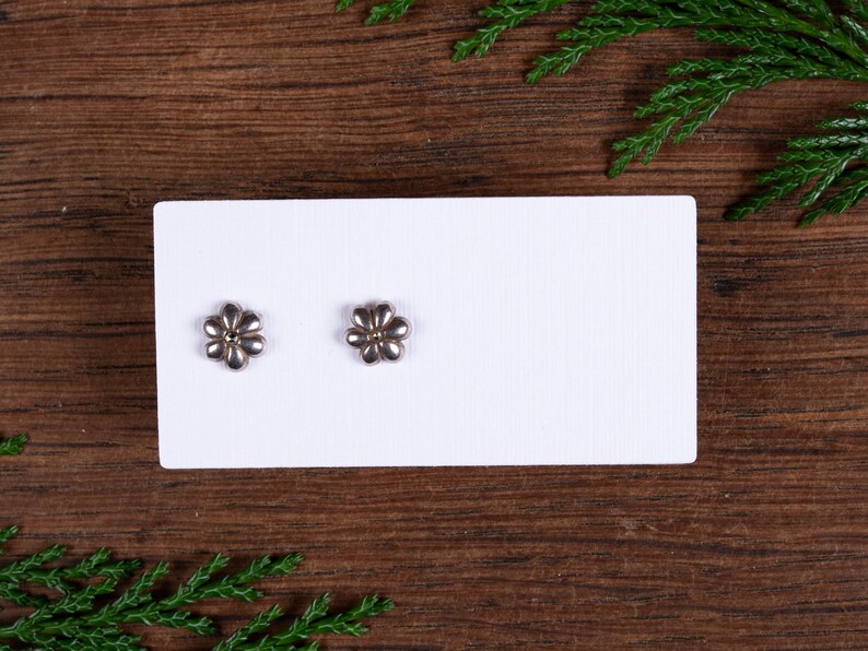 May include: A pair of flower-shaped stud earrings. Each earring has a silver-coloured, six-petal design with a gold-coloured centre. The earrings are displayed on a white card against a dark wood background.