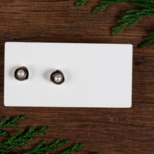 May include: A pair of silver stud earrings, each with a small, round, white pearl set within a textured, petal-like design. The earrings are displayed on a white card against a dark wood background, with green foliage accents.
