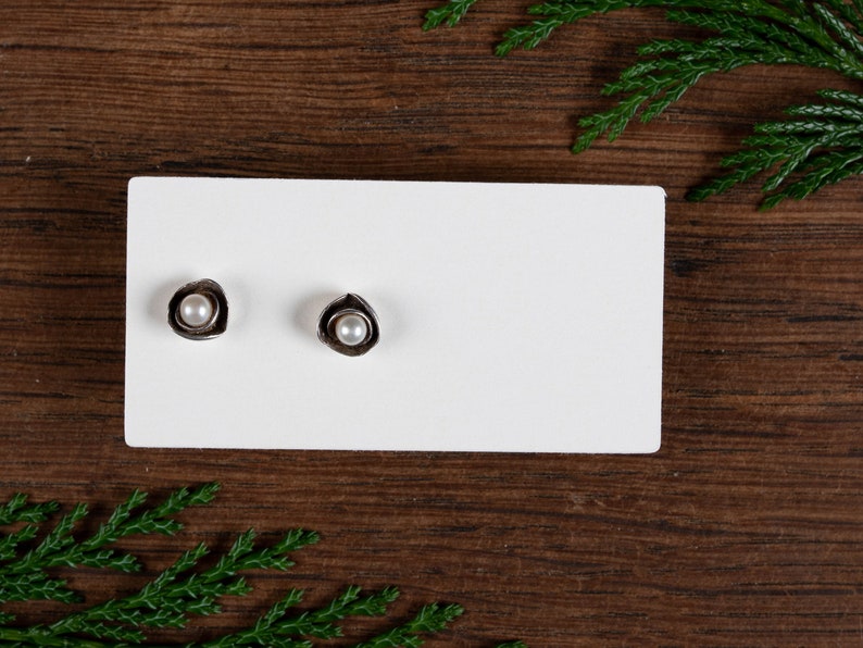 May include: A pair of silver stud earrings, each with a small, round, white pearl set within a textured, petal-like design. The earrings are displayed on a white card against a dark wood background, with green foliage accents.