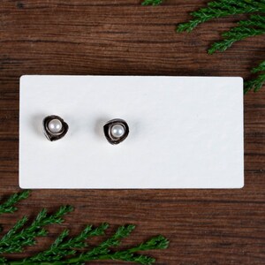 May include: Two silver stud earrings, each with a small, round, white pearl set in an irregular, textured metal frame. The earrings are presented on a white rectangular card against a dark wood background.