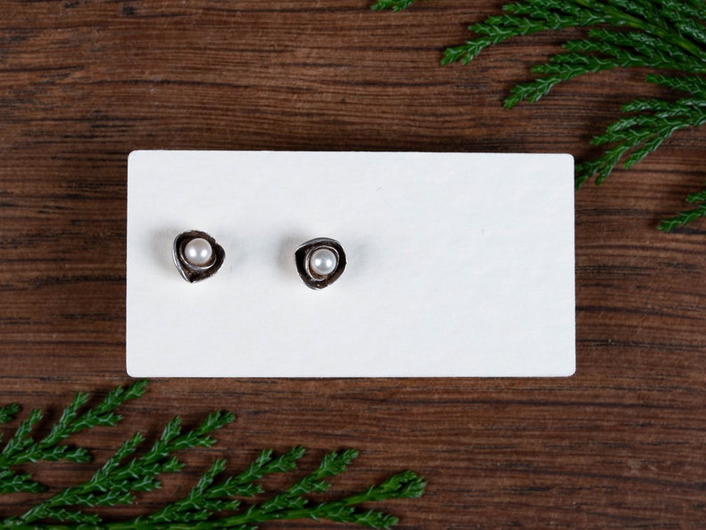 May include: Two silver stud earrings, each with a small, round, white pearl set in an irregular, textured metal frame. The earrings are presented on a white rectangular card against a dark wood background.