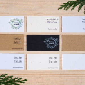 May include: A selection of earring display cards in white, kraft, and black. Some cards display logos and text, including "Fernery Designs" and "Lyme Bay Jewellery". The cards are arranged on a wooden surface.