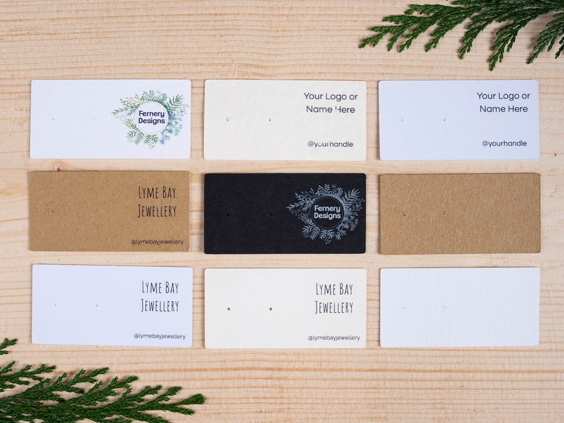 May include: A selection of earring display cards in white, kraft, and black. Some cards display logos and text, including "Fernery Designs" and "Lyme Bay Jewellery". The cards are arranged on a wooden surface.