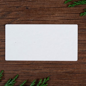 May include: A plain white earring display card with two small holes, set against a dark wood grain background. Green sprigs are in the corners. The card is blank, ready for product display.