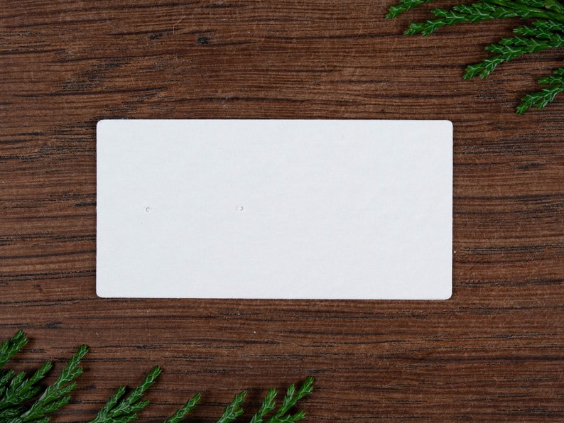 May include: A plain white earring display card with two small holes, set against a dark wood grain background. Green sprigs are in the corners. The card is blank, ready for product display.