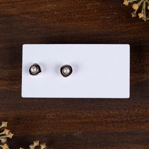 May include: A pair of stud earrings with a silver-coloured, organic-shaped setting. Each earring features a small, round, white pearl in the centre. The earrings are displayed on a white rectangular card against a dark wood background.