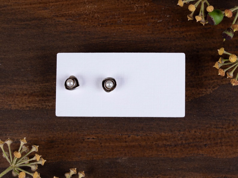 May include: A pair of stud earrings with a silver-coloured, organic-shaped setting. Each earring features a small, round, white pearl in the centre. The earrings are displayed on a white rectangular card against a dark wood background.
