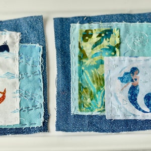 May include: Two denim squares with fabric patches sewn on top. The left square has a white patch with a blue whale and a red fish, and a blue patch with white stitching. The right square has a green and blue floral patch, a light blue patch with white stitching, and a white patch with a blue mermaid.