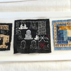 May include: Three fabric squares with different designs. The first square is black with a beige border and features a black cat and a floral pattern. The second square is black with a beige border and features a repeating pattern of white figures. The third square is blue with a beige border and features a layered design of blue and beige fabric.
