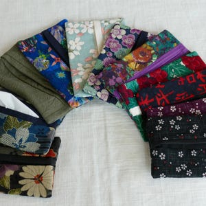 May include: A collection of fabric tissue holders in various floral and patterned designs. The holders feature a range of colors, including blue, green, purple, and black, with visible zippers and openings for tissues. The arrangement is on a white surface.