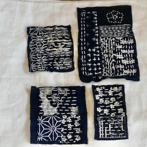 May include: Four small squares of fabric with different patterns and stitching. The fabric is a dark blue color with white stitching. The squares are arranged in a grid pattern.