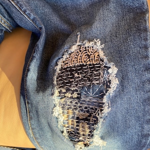 Peut inclure: Gros plan sur une paire de jeans bleus avec un grand patch cousu de mani&egrave;re complexe recouvrant un trou au niveau du genou. Le patch est compos&eacute; de plusieurs tissus diff&eacute;rents, chacun avec un motif et une texture uniques. Les coutures sont r&eacute;alis&eacute;es dans une vari&eacute;t&eacute; de couleurs, notamment le blanc, le noir et le marron.
