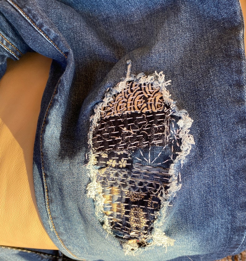 Peut inclure: Gros plan sur une paire de jeans bleus avec un grand patch cousu de mani&egrave;re complexe recouvrant un trou au niveau du genou. Le patch est compos&eacute; de plusieurs tissus diff&eacute;rents, chacun avec un motif et une texture uniques. Les coutures sont r&eacute;alis&eacute;es dans une vari&eacute;t&eacute; de couleurs, notamment le blanc, le noir et le marron.