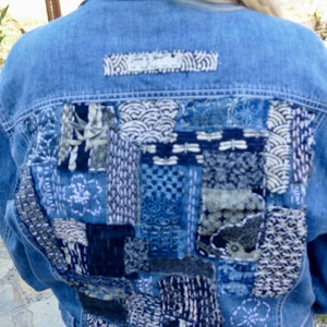 May include: A blue denim jacket with a large patchwork design on the back. The patchwork is made up of different shades of blue and white fabric, and features intricate embroidery.