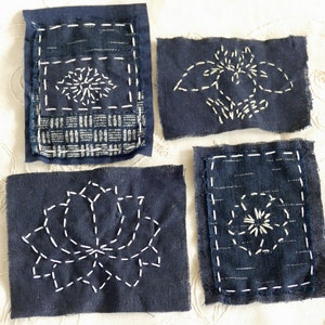 May include: Four blue fabric squares with white embroidery. Each square features a different design, including a flower, a geometric pattern, and two abstract designs.