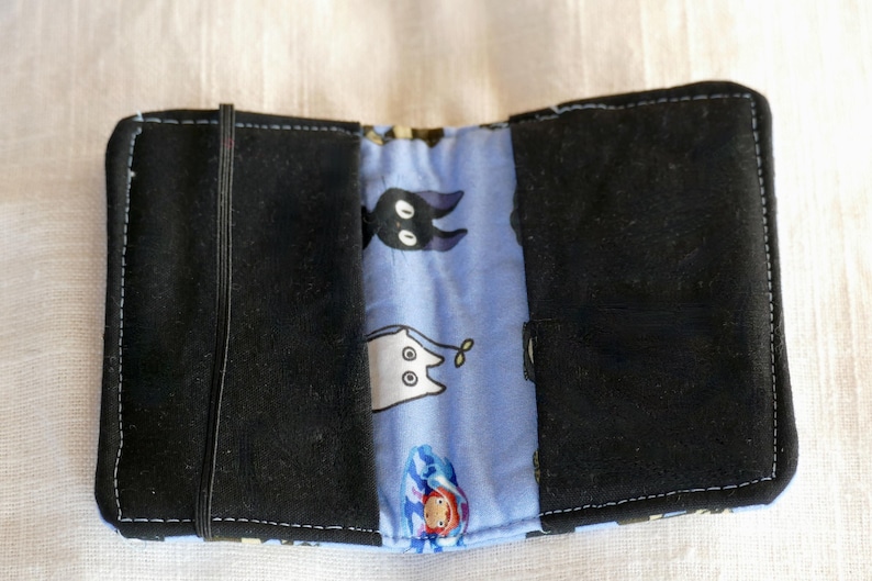 May include: A black fabric wallet with a blue and white fabric lining. The lining has a pattern of cartoon cats and a black cat with white eyes.
