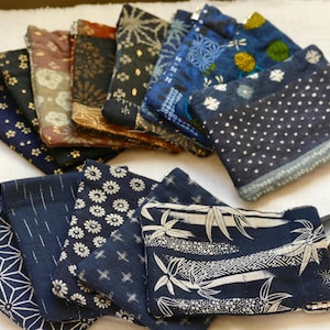 May include: A collection of small, rectangular fabric pouches featuring various indigo and dark-toned patterns, including floral, geometric, and bamboo motifs. Each pouch is zippered and designed to hold small items like coins and cards.
