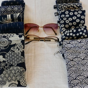May include: A collection of rectangular fabric pouches featuring various dark blue and white Japanese-inspired geometric and floral patterns, arranged in two vertical columns with two pairs of eyeglasses placed horizontally in the center.