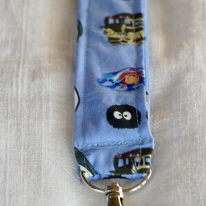 May include: A blue fabric keychain with a repeating pattern of cartoon characters from the movie "My Neighbor Totoro". The keychain has a silver metal clasp.