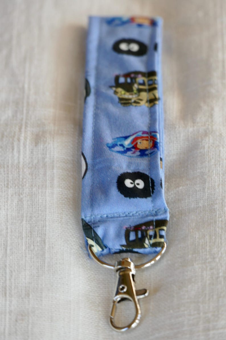 May include: A blue fabric keychain with a repeating pattern of cartoon characters from the movie "My Neighbor Totoro". The keychain has a silver metal clasp.