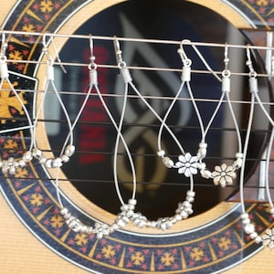 May include: Silver dangle earrings with a flower charm and silver beads. The earrings are hanging on a guitar.
