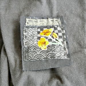 Peut inclure: Un patch en tissu noir avec un motif floral. Le patch est fait de diff&eacute;rents tissus avec divers motifs, dont un motif &agrave; carreaux, un motif &agrave; vagues et un motif g&eacute;om&eacute;trique. Les fleurs sont jaunes et les tiges sont vertes.