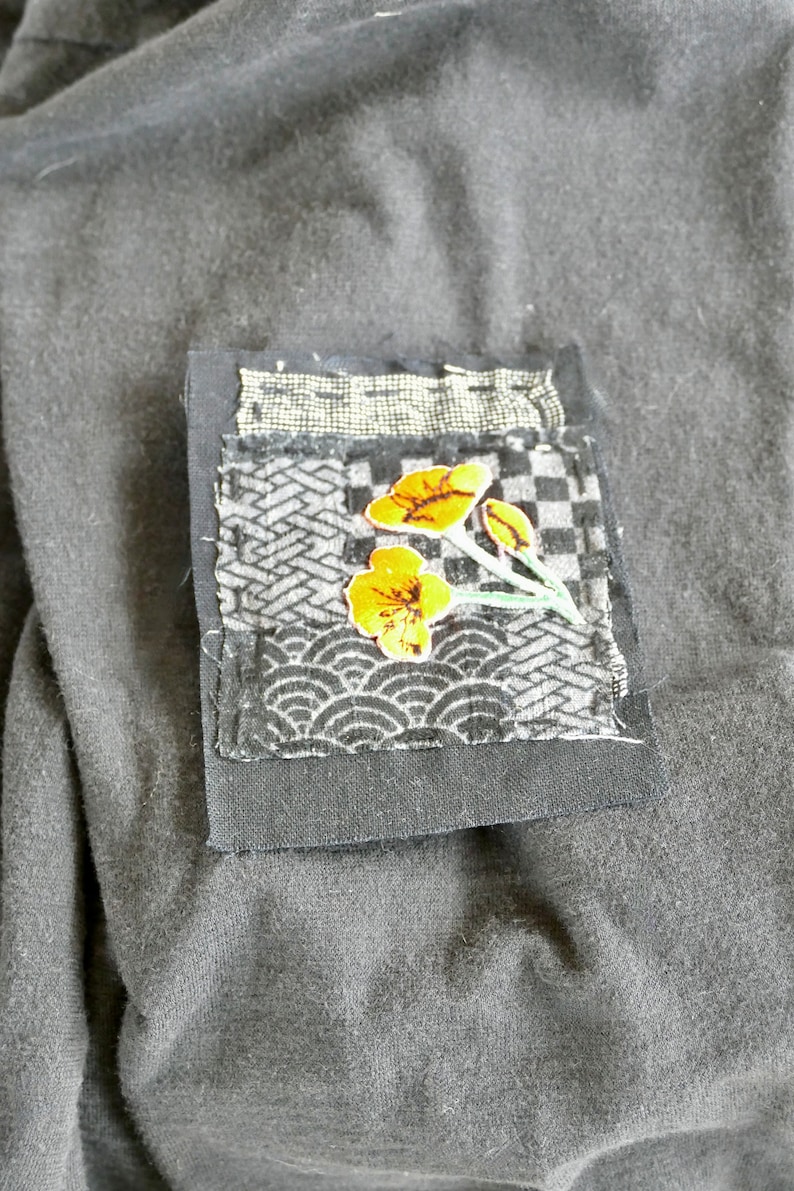 Peut inclure: Un patch en tissu noir avec un motif floral. Le patch est fait de diff&eacute;rents tissus avec divers motifs, dont un motif &agrave; carreaux, un motif &agrave; vagues et un motif g&eacute;om&eacute;trique. Les fleurs sont jaunes et les tiges sont vertes.