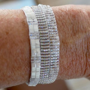 May include: Close-up of three white beaded bracelets stacked on a wrist. The bracelets are made of small, rectangular beads, creating a shimmering effect. The skin tone is fair, with visible texture.