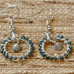 May include: A pair of silver-toned wire-wrapped hoop earrings featuring small, iridescent blue-green beads encircling the frame. Each earring includes a dangling silver-toned seashell charm and a standard hook-style ear wire for attachment.