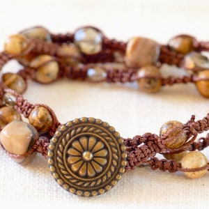 May include: A multi-strand bracelet featuring brown knotted cord woven with smooth, tan, and brown speckled stone beads and translucent gray beads. The bracelet is secured with a round, antique brass-toned metal button closure featuring a floral pattern.