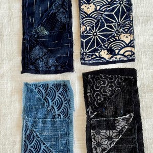 May include: Four rectangular fabric patches featuring various blue and black indigo-dyed patterns. The patches display visible hand-stitched sashiko-style embroidery, frayed edges, and geometric motifs including waves, hemp leaves, and stylized cat faces.