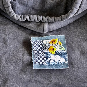 Peut inclure: Un sweat-shirt gris avec un petit patch cousu sur le devant. Le patch est en jean et pr&eacute;sente un motif floral avec des fleurs jaunes et des tiges vertes.