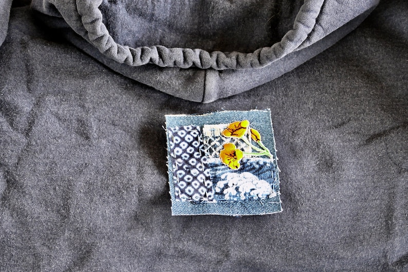Peut inclure: Un sweat-shirt gris avec un petit patch cousu sur le devant. Le patch est en jean et pr&eacute;sente un motif floral avec des fleurs jaunes et des tiges vertes.